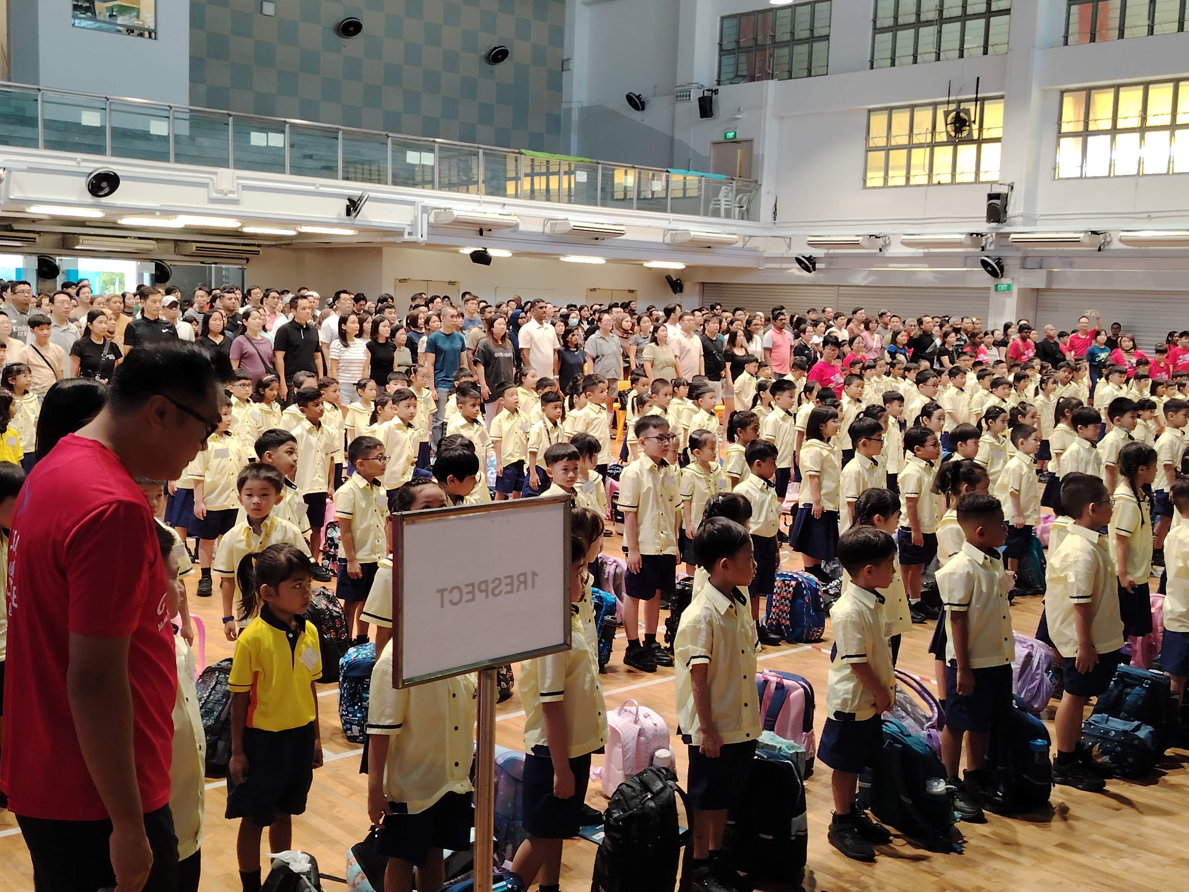 P1 students stand in rows in a school hall for their first morning assembly
